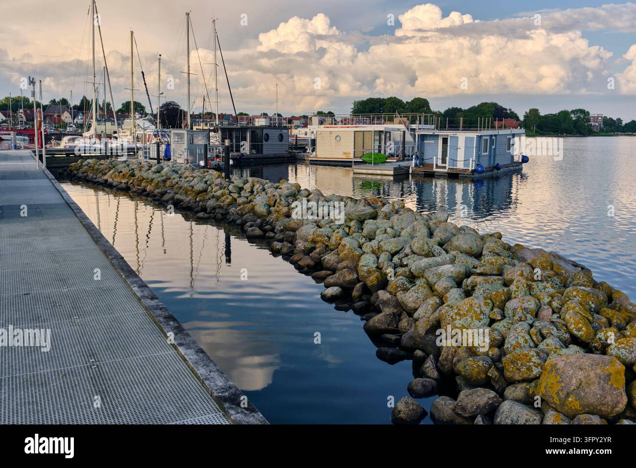Schleswig, Allemagne - 1er juillet 2026 : péniches et port de plaisance de Schleswig, Allemagne, derrière une digue, maisons flottantes et voiliers amarrés dans un port calme lors d'une soirée d'été. Brise-lames en pierre menant à un Banque D'Images