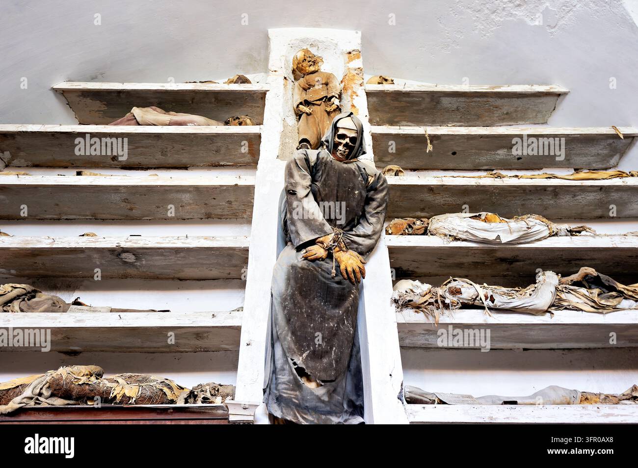 Restes momifiés sur des étagères en bois et un corps squelettique dans une robe de moine aux mains liées dans une crypte funéraire historique en Sicile, Italie Banque D'Images