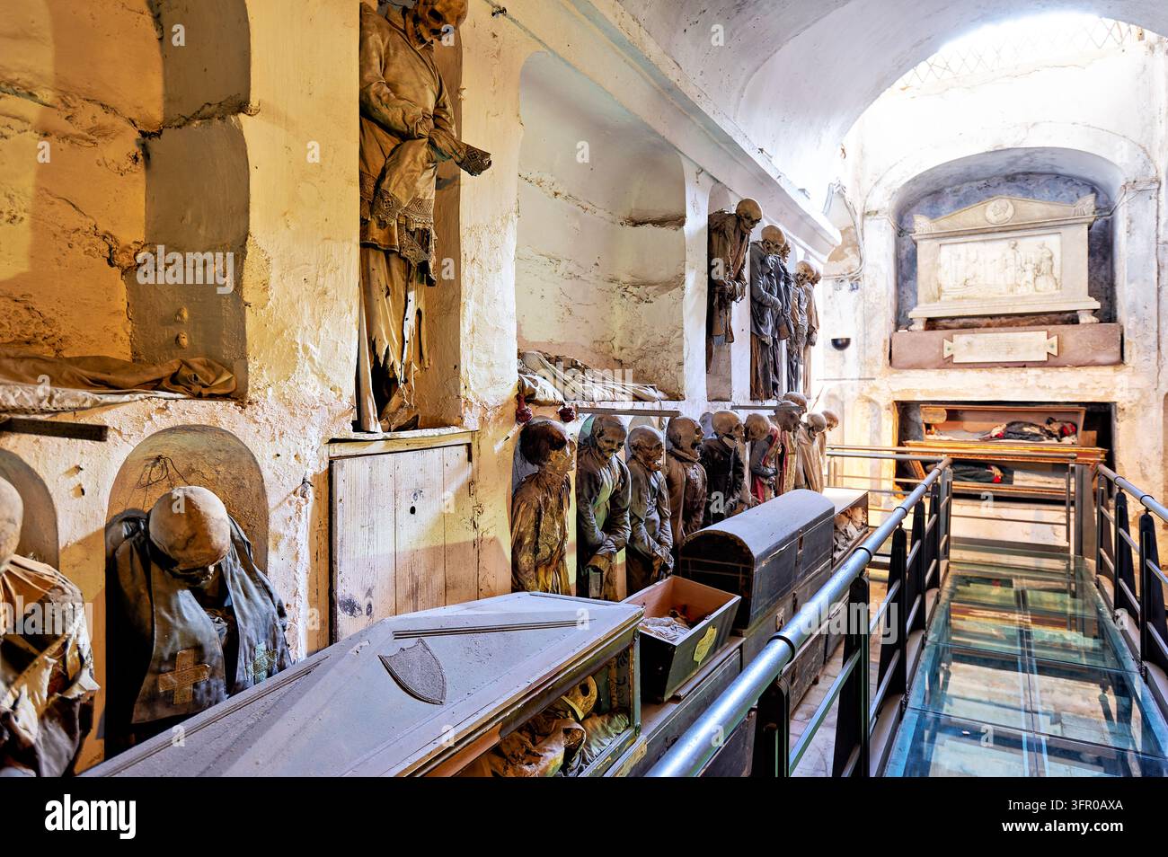 Des corps momifiés vêtus se dressent dans des niches murales au-dessus des cercueils le long d'un couloir voûté avec passerelle en verre dans les catacombes des Capucins, Palerme, Italie. Banque D'Images