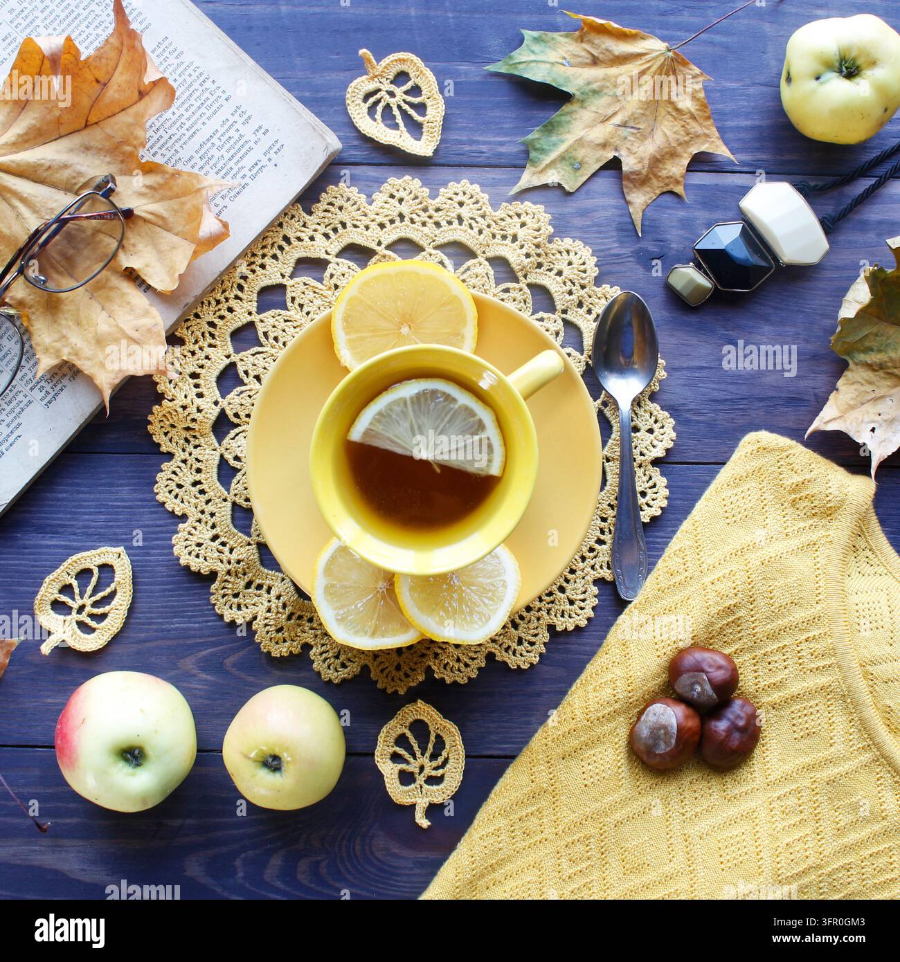 Composition automnale douillette avec une tasse de thé jaune au citron, une napperon en dentelle au crochet et des feuilles faites à la main, un livre ouvert, des pommes et des châtaignes sur du bois sombre Banque D'Images