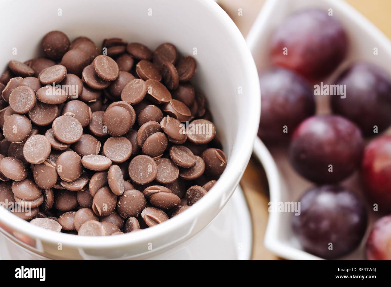 Chips de chocolat au lait dans un bol blanc avec des prunes fraîches en arrière-plan. Ingrédients de boulangerie pour des desserts maison, des recettes sucrées et des préparations de confiserie Banque D'Images