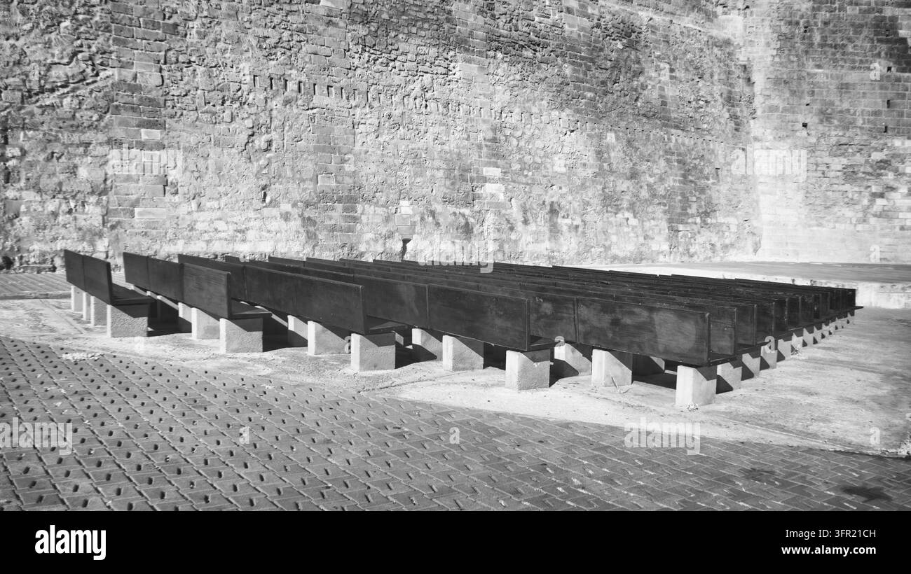Des rangées de bancs en bois sombre et vides avec des supports en béton sont disposées sur une surface pavée, devant un mur de briques vieilli et texturé à Majorque, Espagne Banque D'Images