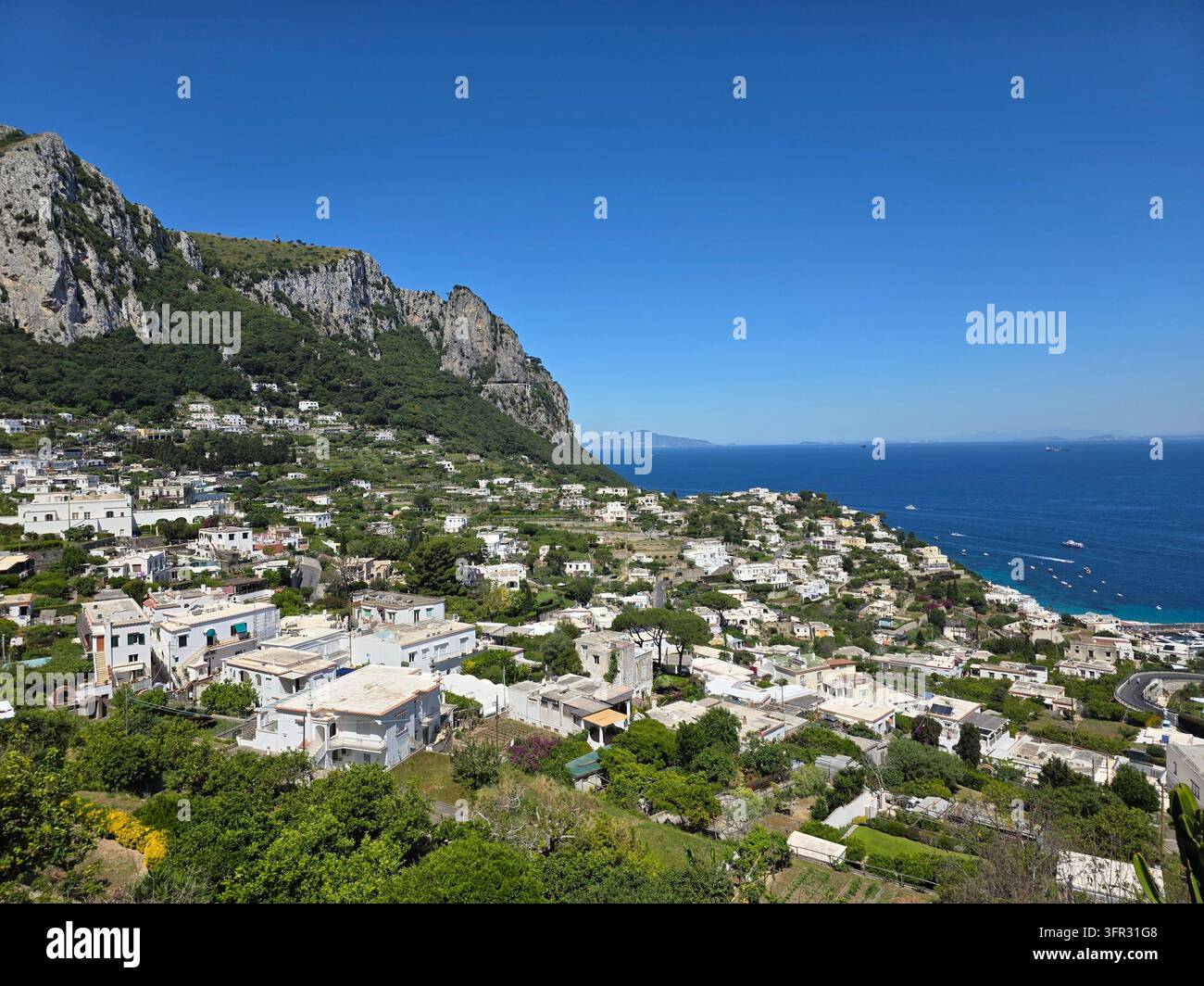 Une montagne rocheuse escarpée s'élève au-dessus de maisons blanches en terrasses qui dégringolent vers une mer calme et d'un bleu profond à Capri. Un panorama clair de fin de matinée de l'île - Image de stock capturée avec un smartphone