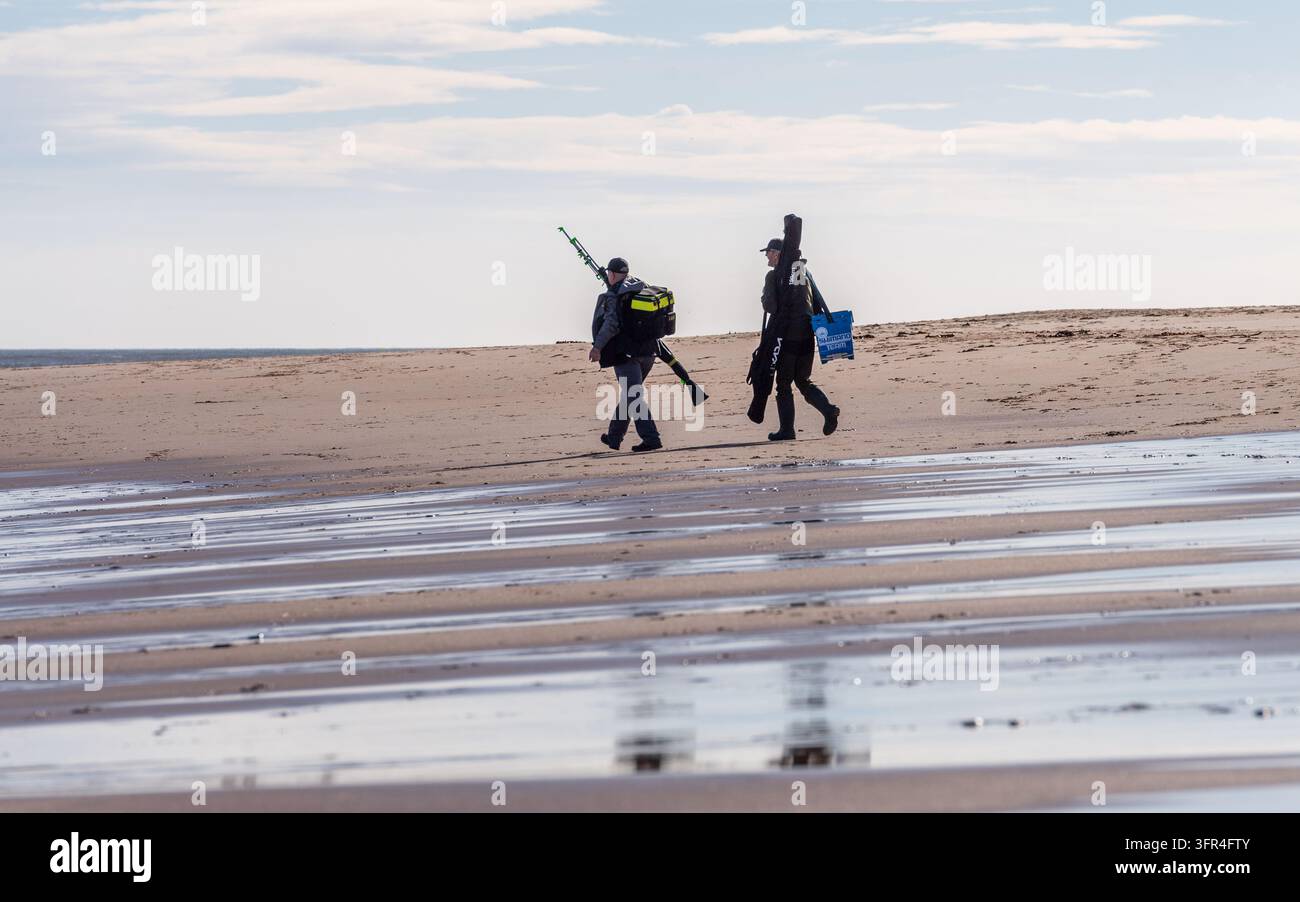 Deux pêcheurs marchant le long d'une plage de sable portant du matériel et de l'équipement de pêche pour une journée de pêche à Druridge Bay, Northumberland, Angleterre. Banque D'Images