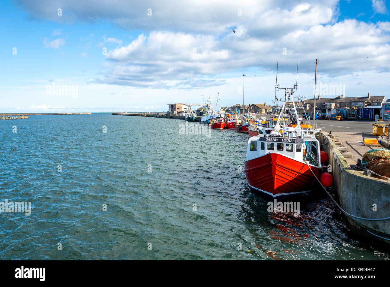 Un chalutier rouge vif repose le long d'un quai en pierre dans un port maritime animé avec plusieurs autres navires amarrés dans la ville de pêche d'Amble. Banque D'Images