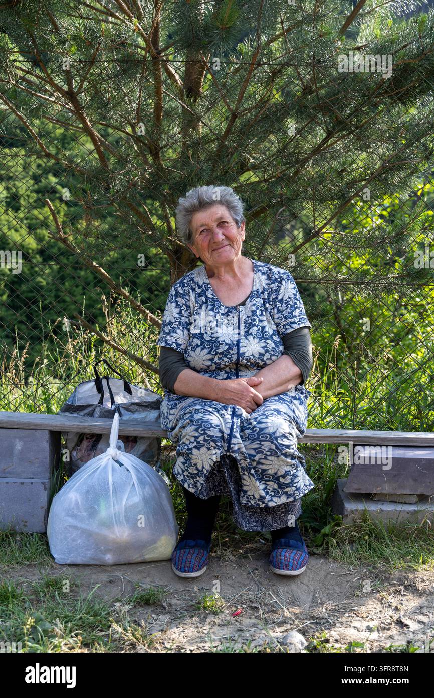 Ancienne femme assise sur un banc en bois avec des sacs de courses en plastique, province de Lori, Haghpat, Arménie Banque D'Images