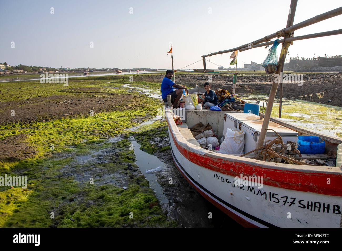 Deux hommes sont assis sur un petit bateau de pêche échoué sur un lit de crique couvert d'algues boueuses à marée basse, Gujarat, Mandvi, Inde Banque D'Images