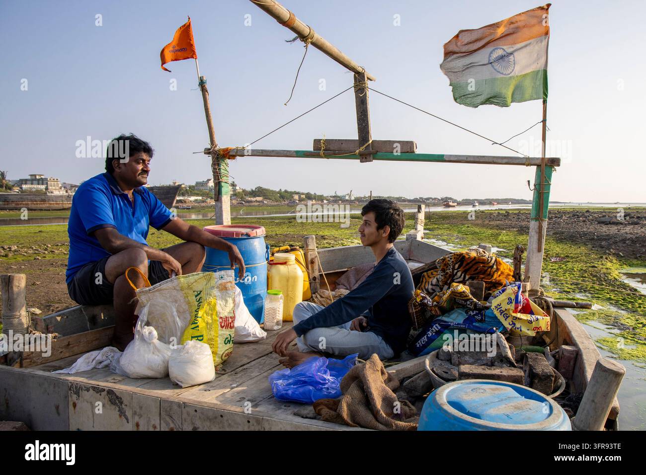 Deux hommes discutent assis sur un petit bateau de pêche décoré de drapeaux sur un lit de crique boueux à marée basse, Gujarat, Mandvi, Inde Banque D'Images