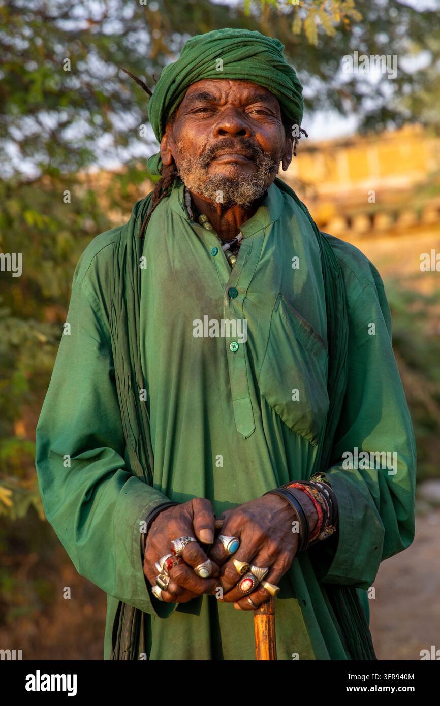 Un homme âgé portant un turban vert et une robe verte ornée de nombreux anneaux s'appuie sur une canne en bois, Gujarat, Mandvi, Inde Banque D'Images