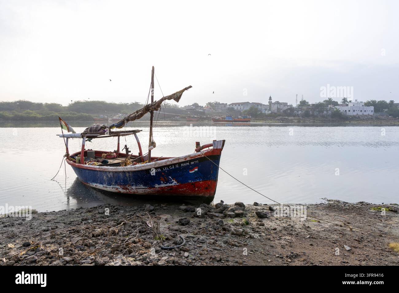 Un bateau de pêche traditionnel en bois peint en bleu et rouge est amarré sur une eau calme près d'une ville côtière, Gujarat, Mandvi, Inde Banque D'Images