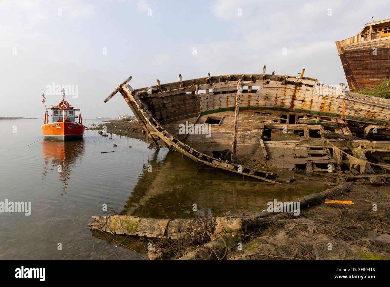 Un petit bateau de pêche flotte à côté de la coque en décrépitude d'un navire en bois abandonné à marée basse, Gujarat, Mandvi, Inde Banque D'Images