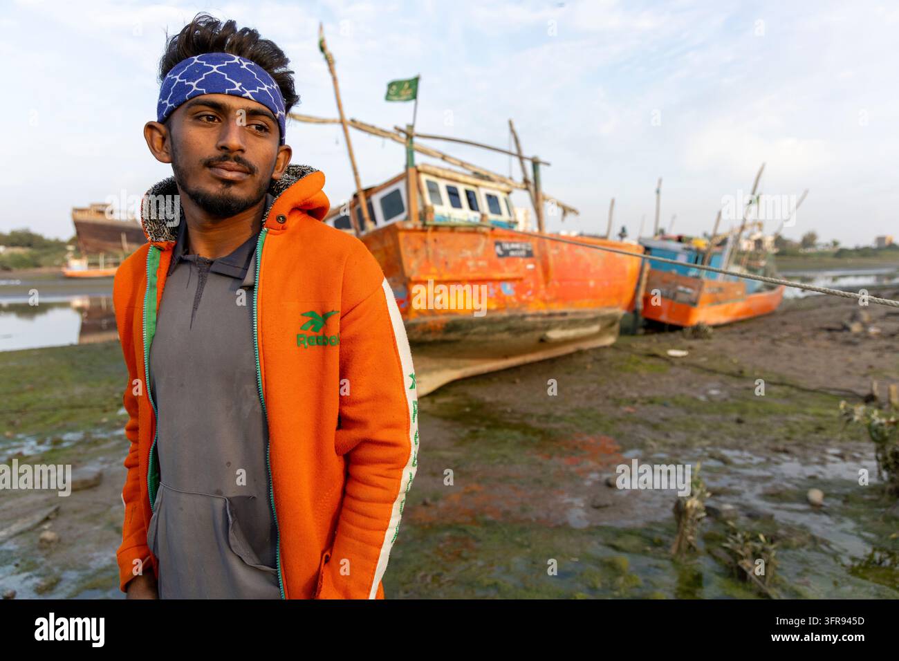 Un jeune homme portant un bandeau à motifs et une veste à capuche se tient près des bateaux de pêche, Gujarat, Mandvi, Inde Banque D'Images