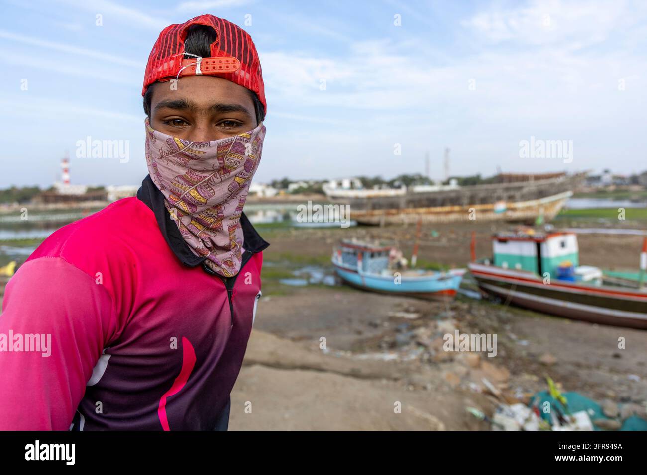 Jeune homme portant une casquette rouge et un couvre-visage à motifs debout près de bateaux de pêche et d'une chantier naval, Gujarat, Mandvi, Inde Banque D'Images