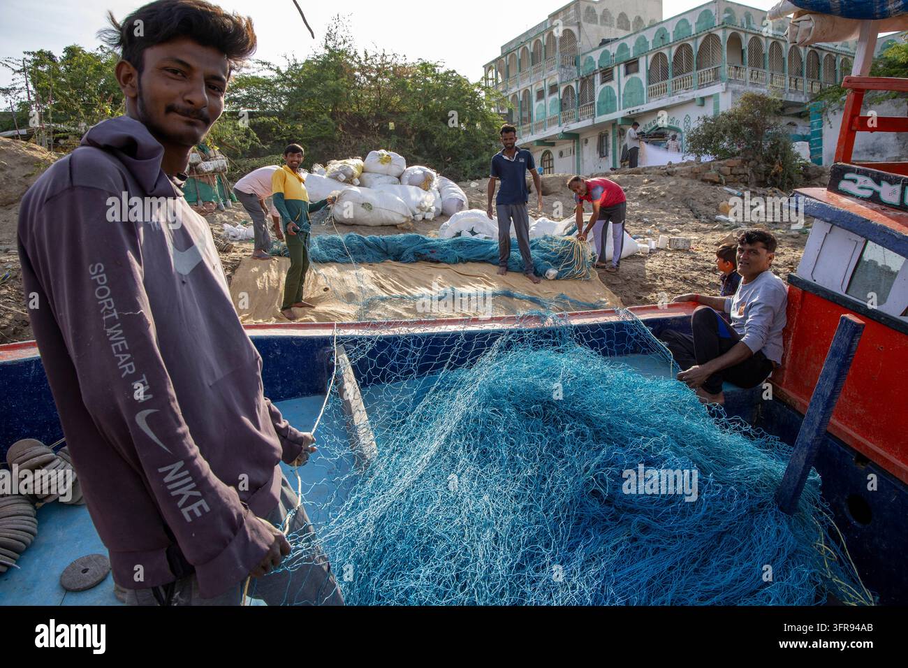 Pêcheur tenant un filet de pêche bleu à bord d'un bateau avec des personnes travaillant à terre à proximité, Gujarat, Mandvi, Inde Banque D'Images
