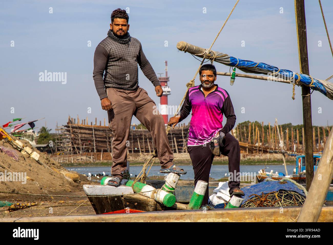 Deux pêcheurs posent sur un bateau avec un phare rayé et une chantier de dhow en arrière-plan, Gujarat, Mandvi, Inde Banque D'Images