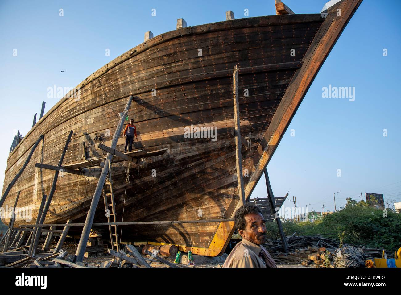 Homme en premier plan en gros plan debout près d'une coque de navire géante en bois avec un ouvrier en équilibre sur une plateforme en bois, Gujarat, Mandvi, Inde Banque D'Images