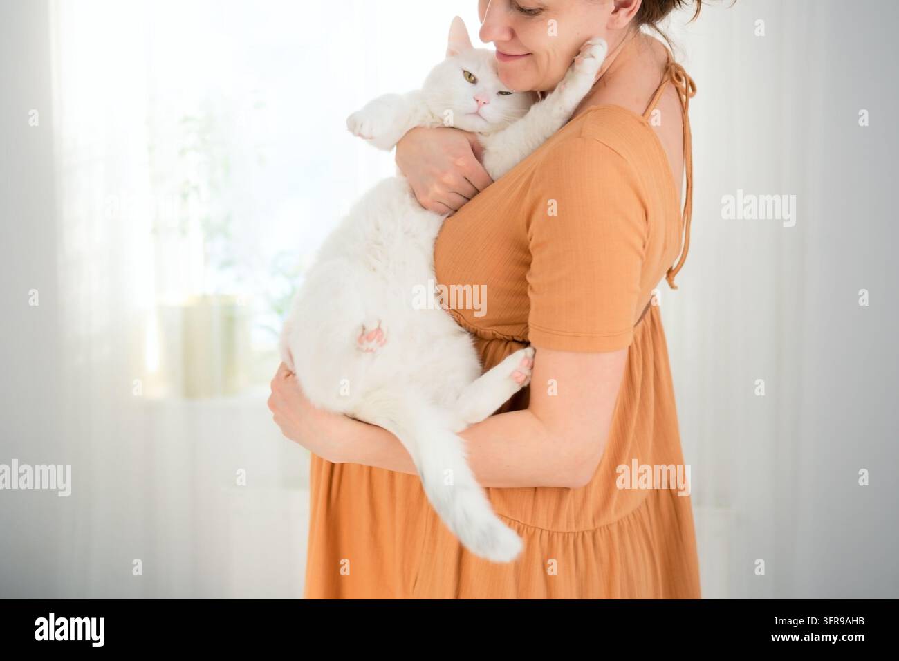Une femme enceinte tient un chat blanc dans ses bras dans une pièce lumineuse. La mère enceinte qui embrasse son animal de compagnie présente un moment prudent et préoccupé concernant toutes Banque D'Images