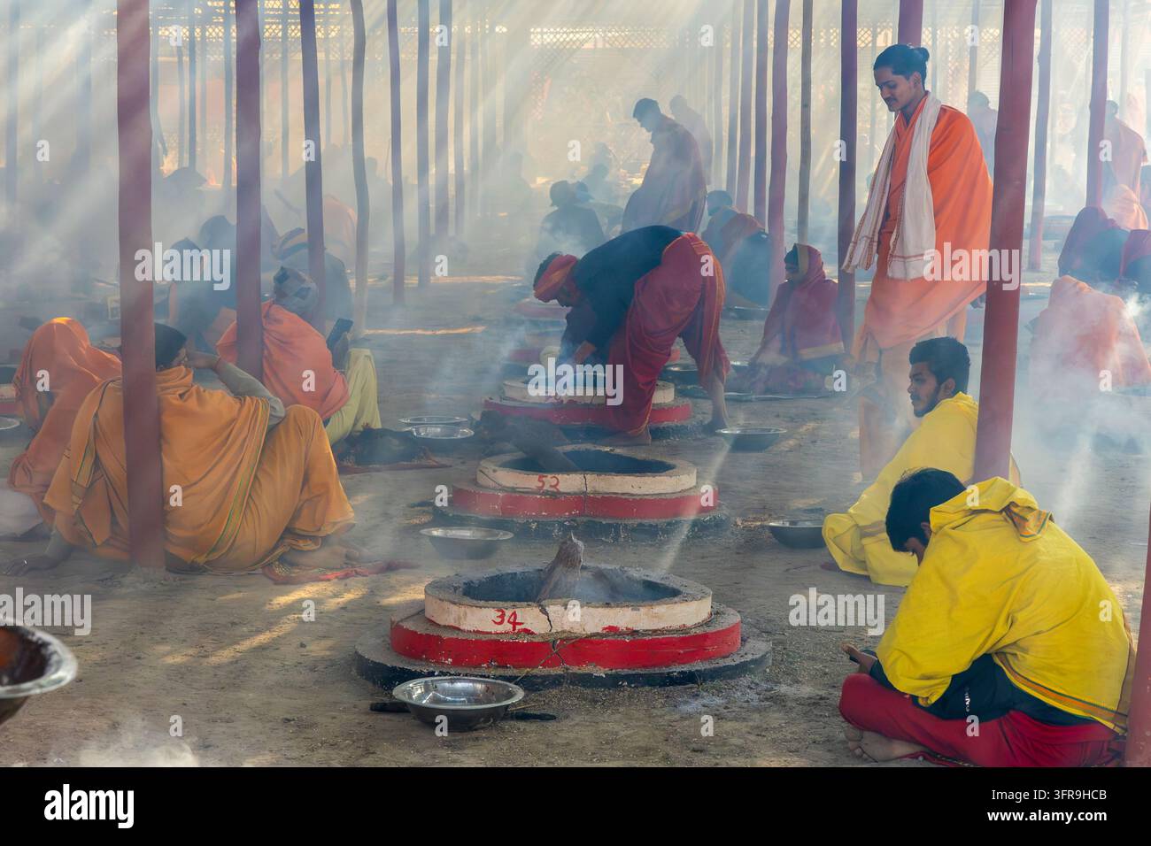 Des dévots en robes orange et rouges entretiennent des foyers fumants sous un abri éclairé par les rayons du soleil lors d’un rituel puja, Uttar Pradesh, Prayagraj, Inde Banque D'Images
