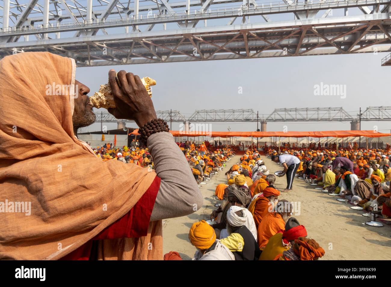 Sadhu en châle orange souffle une conque alors que des milliers de dévots sont assis en rangées pour un repas à la Maha Kumbh Mela, Uttar Pradesh, Prayagraj, Inde Banque D'Images