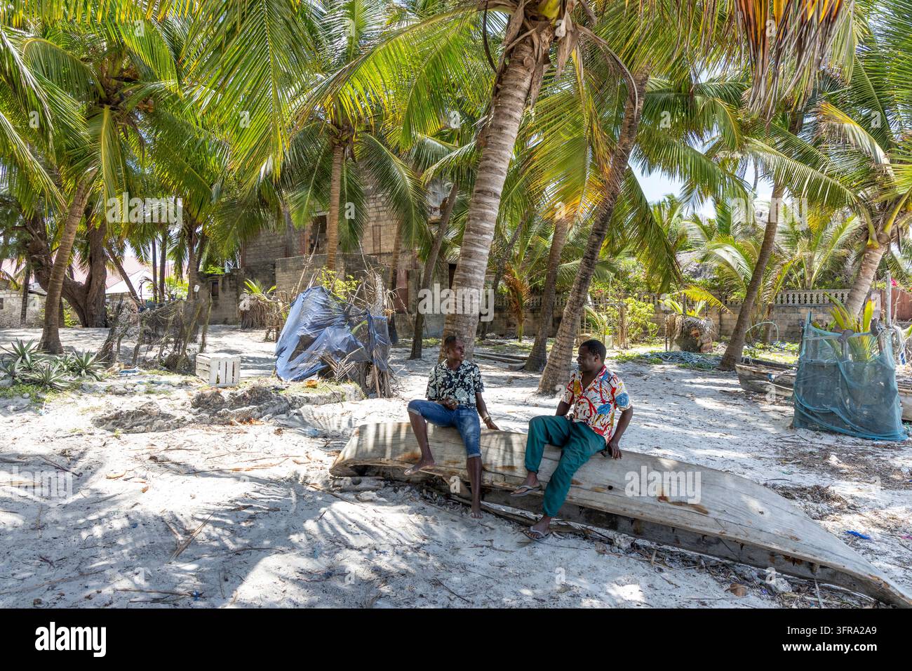 Deux hommes discutant assis sur une vieille coque de pirogue en bois sous des palmiers à cocos sur une plage de sable bordée de filets de pêche et de huttes simples, Zanzibar Banque D'Images