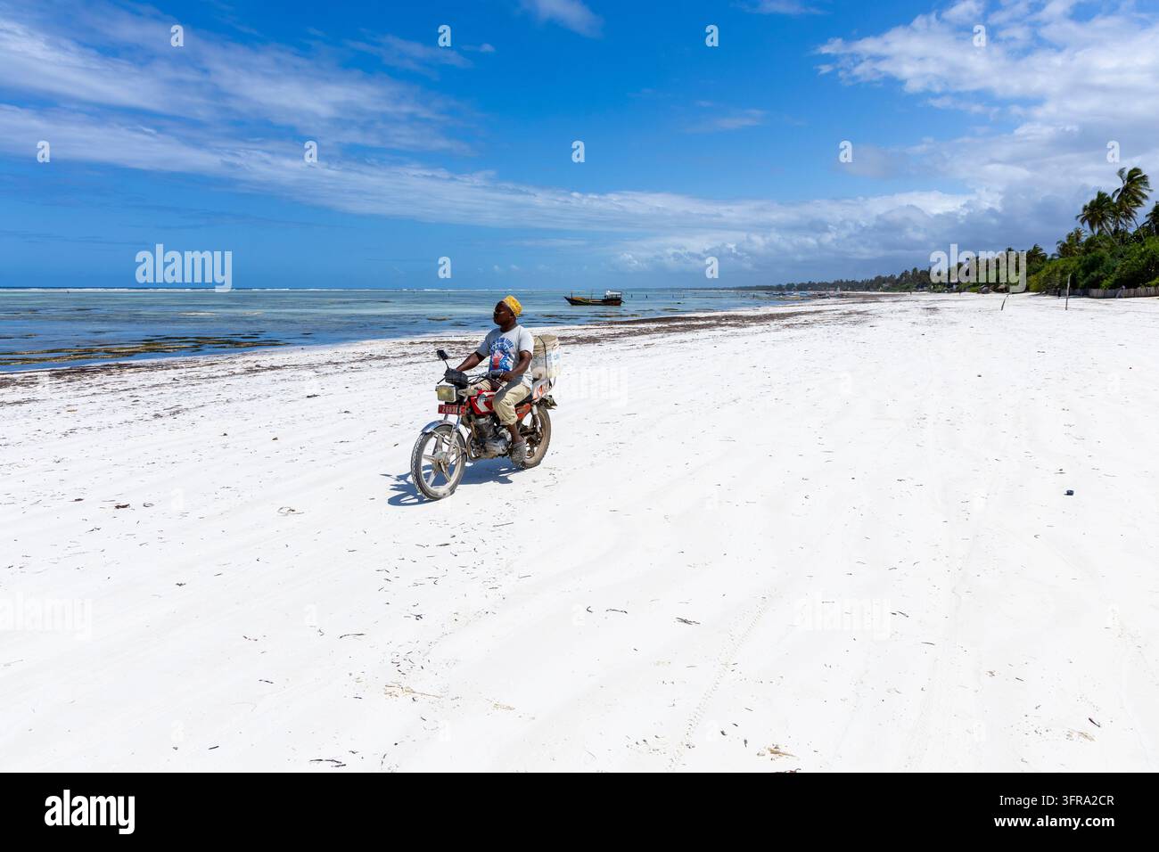 Homme portant une casquette kofia jaune chevauchant une moto rouge le long d'une vaste plage de sable blanc avec un lagon turquoise et un bateau en bois sous un ciel bleu profond Banque D'Images