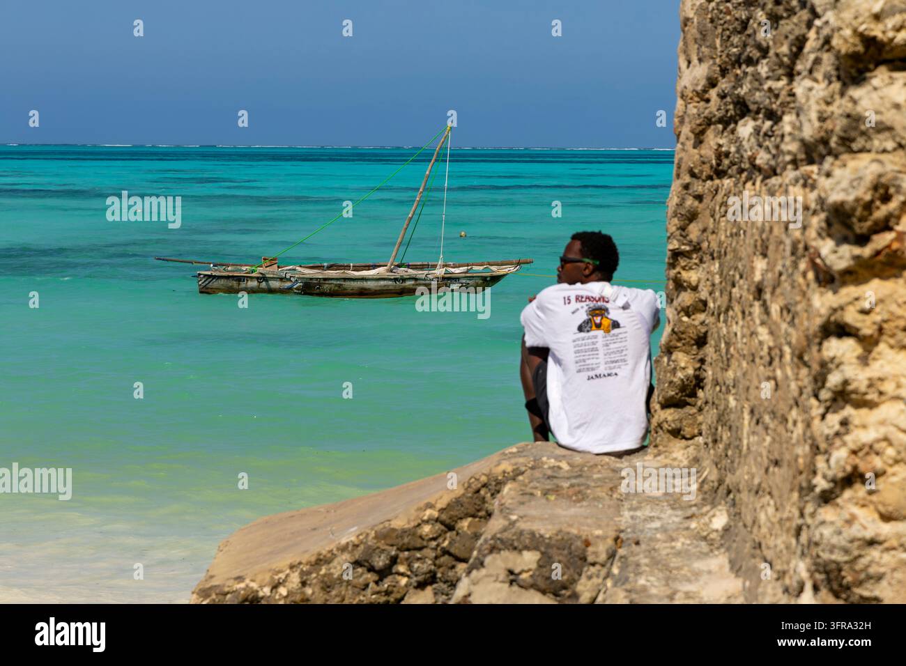 Homme portant un T-shirt blanc de la Jamaïque et des lunettes de soleil, assis sur un mur de pierre coralline et regardant une voile traditionnelle en bois amarrée dans le turquoise Banque D'Images