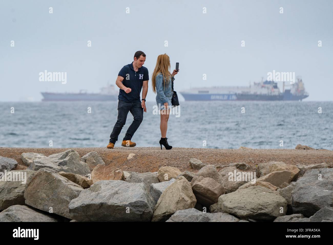 DANSANT POUR LA CAMÉRA AU BORD DE LA MER, PLAYA DE LA BARCELONETA REPORTAGE, BARCELONE, ESPAGNE : Un homme en polo bleu exécute une danse tandis qu'une femme aux longs cheveux blonds le filme avec son smartphone sur une jetée en pierre, avec la mer et des cargos en arrière-plan. INFO : La plage de Barceloneta est la destination balnéaire la plus célèbre et vibrante de Barcelone. Célèbre pour ses sables en pente et sa promenade animée bordée d'arbres, elle allie bains de soleil et sports nautiques avec des bars et restaurants animés en bord de mer, des jetées en béton et des zones d'exercice. Banque D'Images