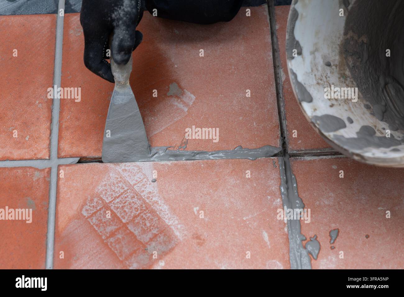 Une main gantée étale du mortier gris le long des joints de carreaux de terre cuite. Banque D'Images