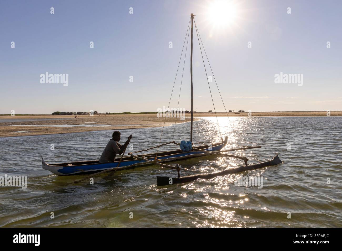Un homme Vezo pagaie une pirogue à balancier en bois sur une eau calme avec un village de pêcheurs visible au loin, Menabe, Belo sur Mer, Madagascar Banque D'Images