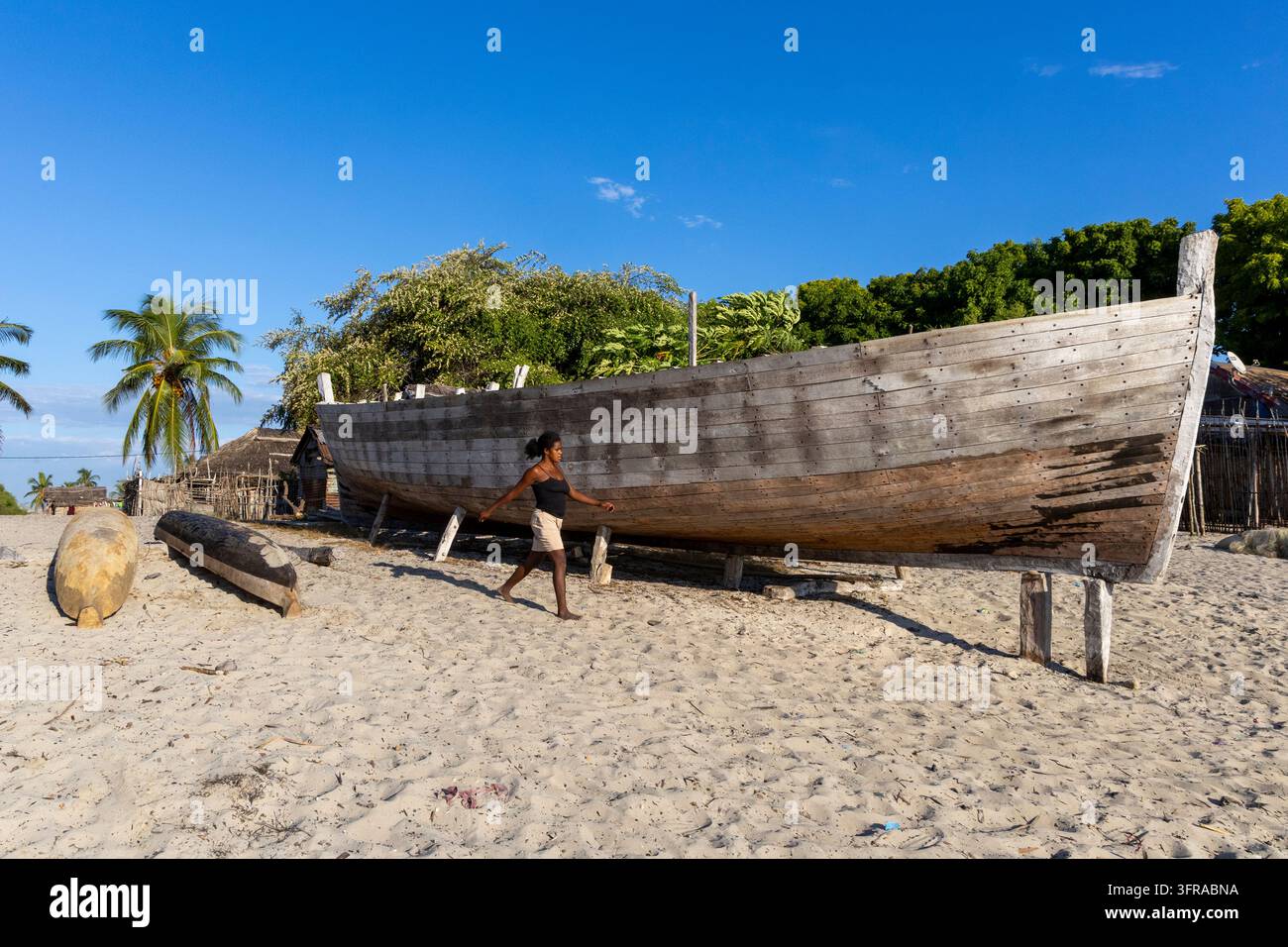 Un grand vieux bateau en bois repose sur des pilotis en bois sur la plage tandis qu'une femme Vezo passe à proximité des huttes, Menabe, Belo sur Mer, Madagascar Banque D'Images