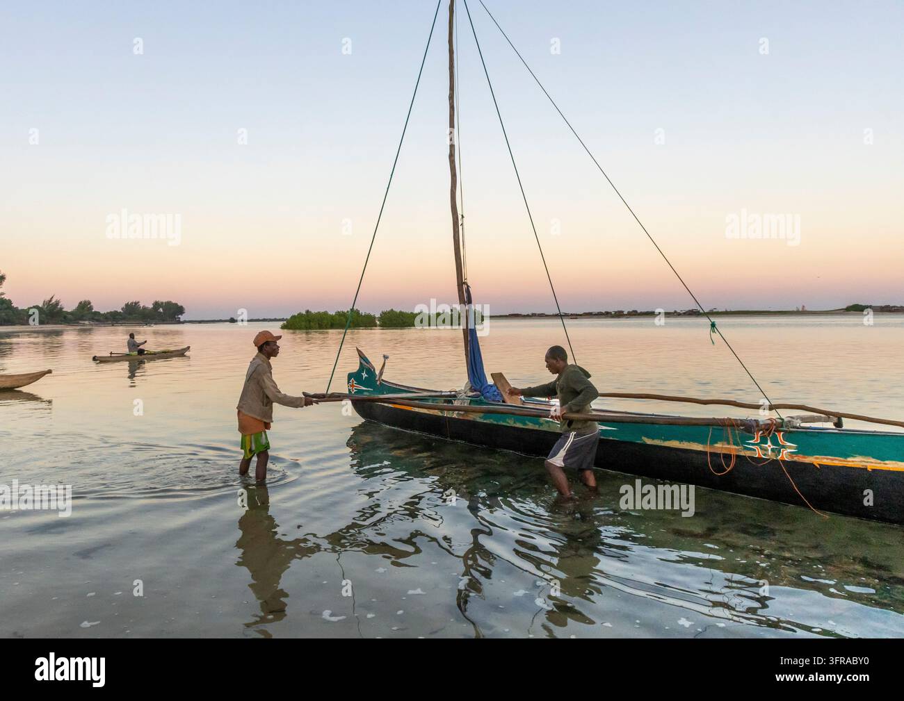 Deux pêcheurs Vezo pataugent dans des eaux peu profondes tout en préparant une pirogue à balancier pour le départ au coucher du soleil, tandis qu'un autre pagaye en arrière-plan, Mena Banque D'Images