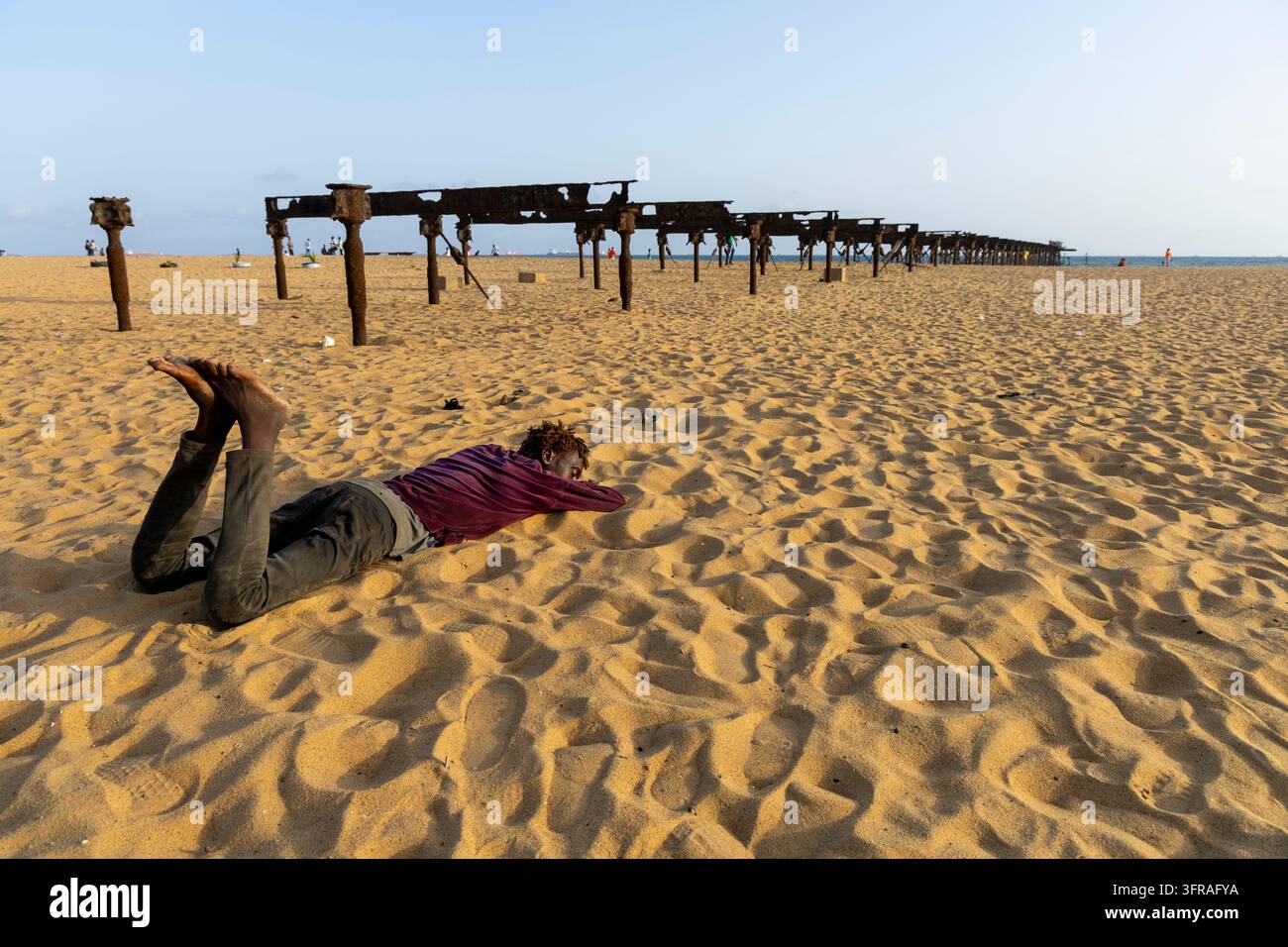 Homme allongé sur le sable près des restes rouillés d'un vieux quai s'étendant dans l'océan, Région Maritime, Lomé, Togo Banque D'Images