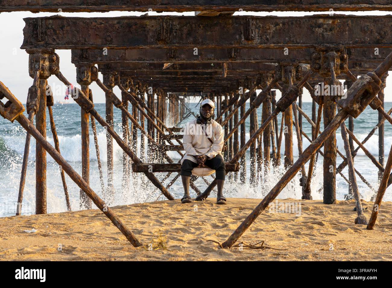 Homme assis sur une poutre sous la structure rouillée d'un vieux pier en fer au bord de l'océan, région maritime, Lomé, Togo Banque D'Images