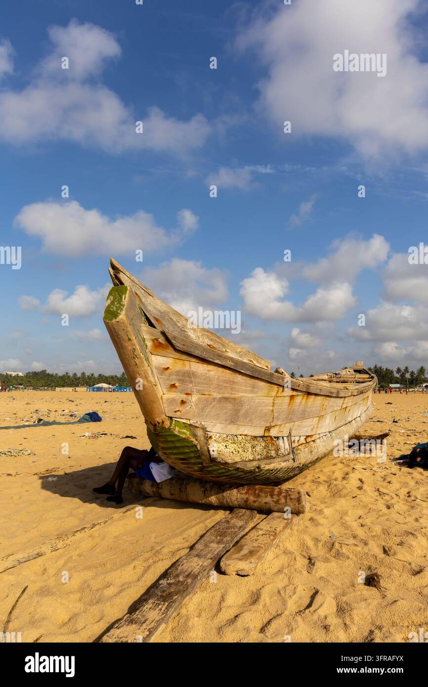 Bateau de pêche traditionnel en bois reposant sur le sable avec une personne abritée sous sa coque, Région Maritime, Lomé, Togo Banque D'Images