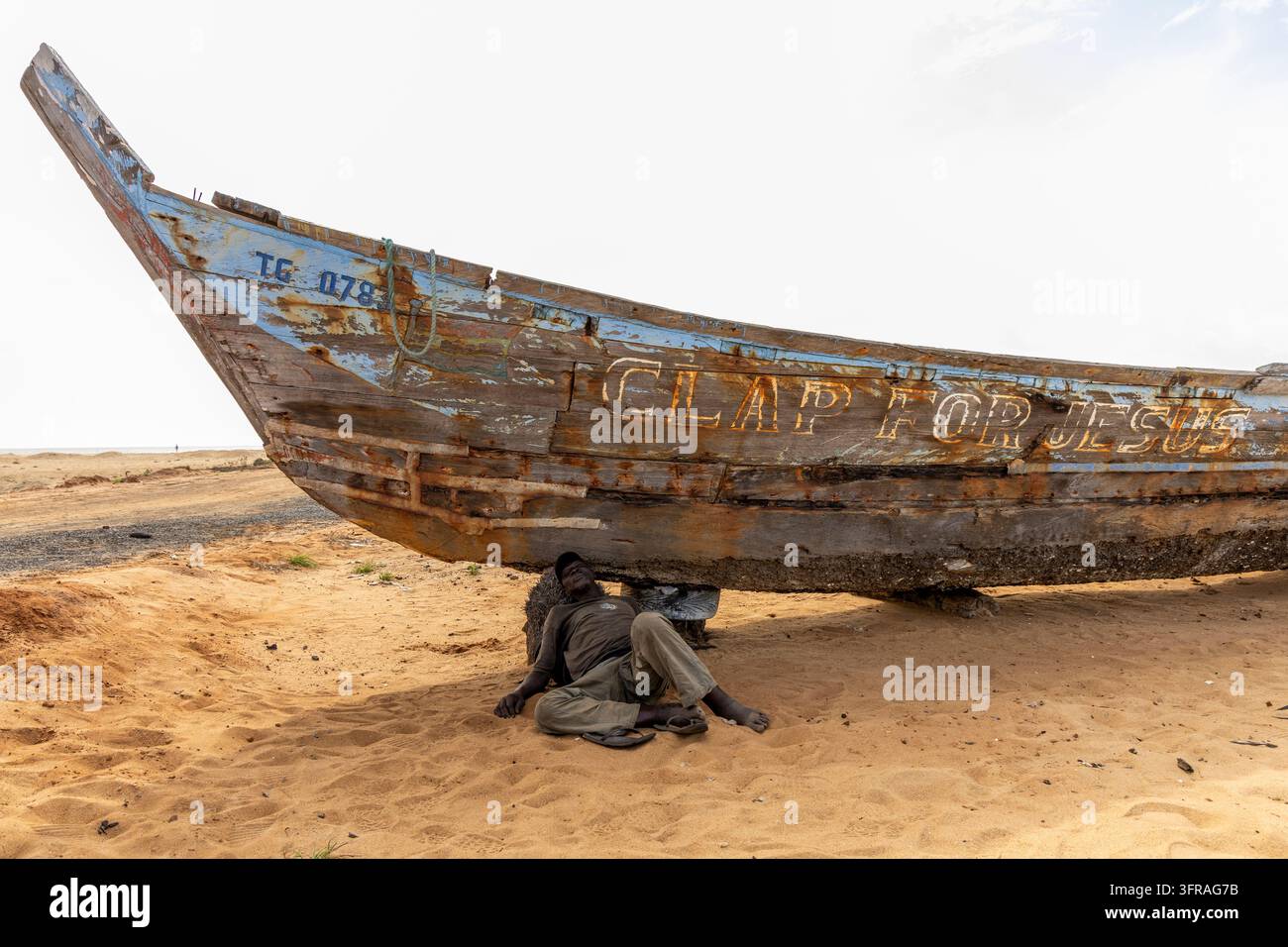 Un homme sommeille à l'ombre sous un bateau de pêche en bois patiné peint Clap for Jesus, Région Maritime, Lomé, Togo Banque D'Images