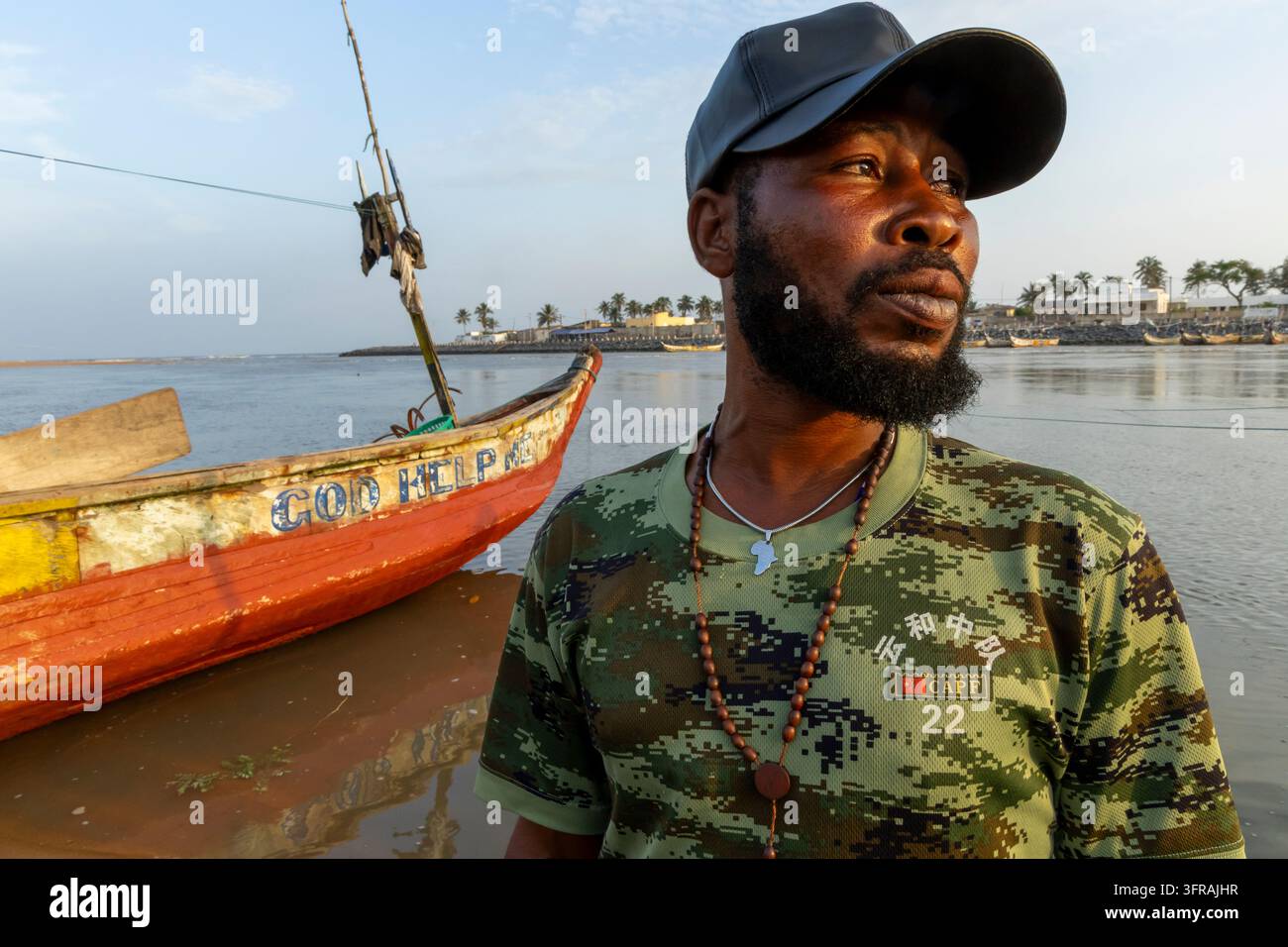 Pêcheur portant une casquette debout à côté d'une pirogue en bois peinte, Région Maritime, Aneho, Togo Banque D'Images