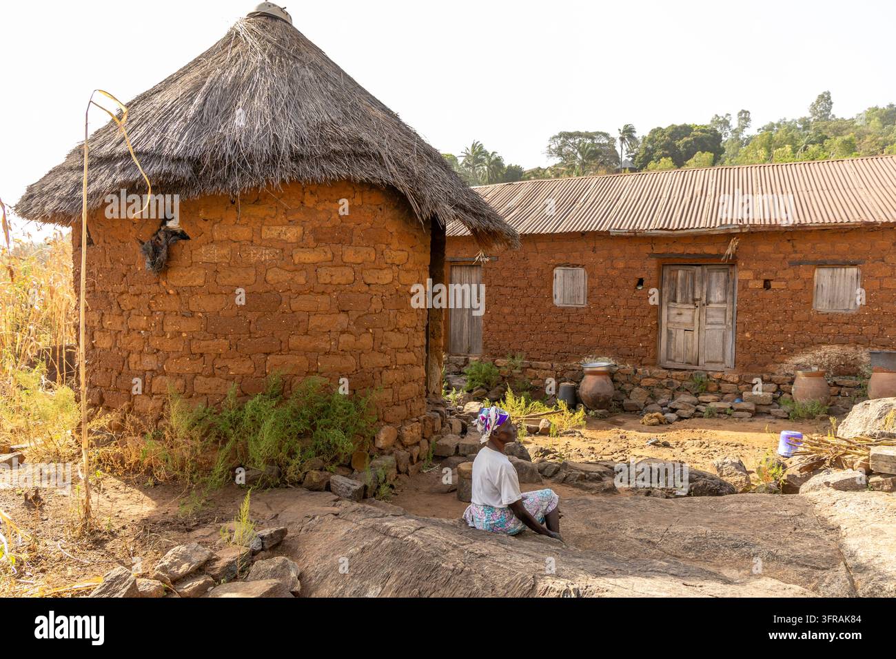 Une maison traditionnelle ronde en briques de boue avec un toit conique en chaume se dresse dans un complexe rural tandis qu'une femme est assise à proximité, région de Kara, mont Kabyé, Togo Banque D'Images