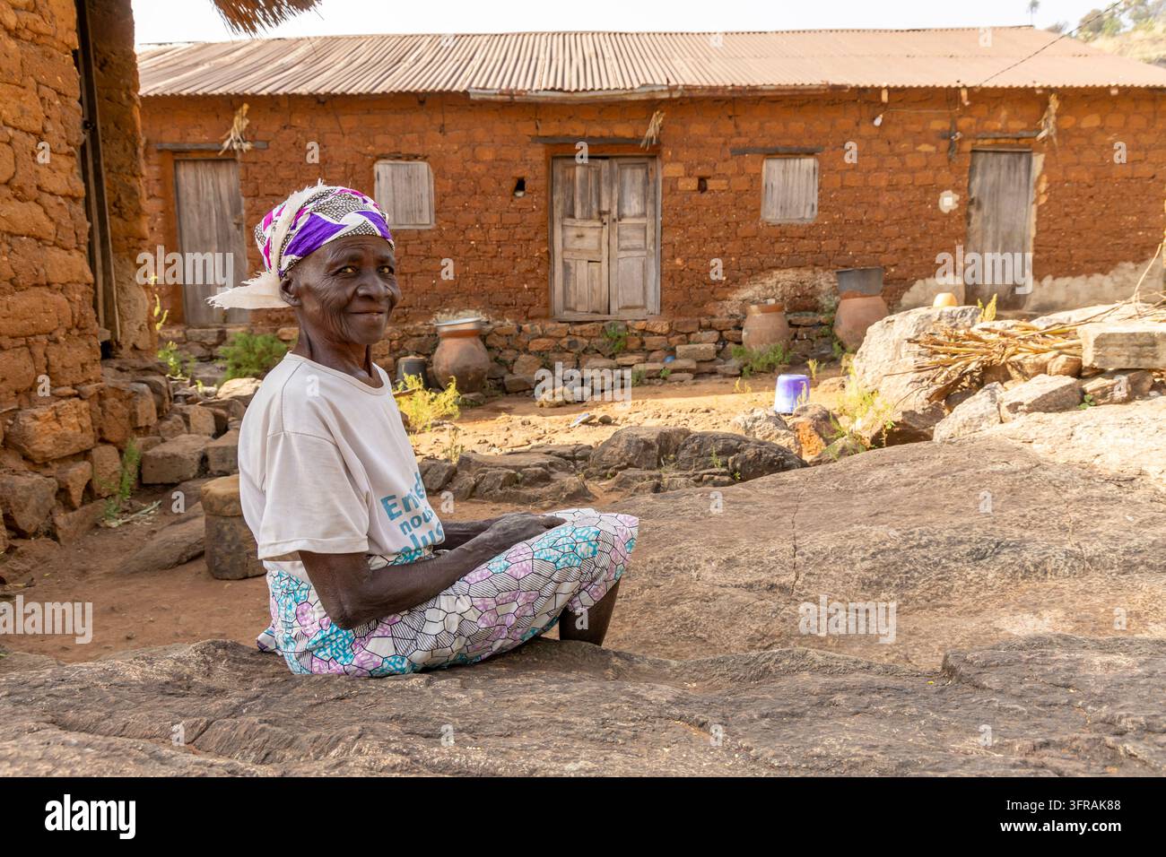 Une femme âgée est assise devant une maison en briques de boue, portant une jupe enveloppante à motifs et un foulard, région de Kara, mont Kabye, Togo Banque D'Images