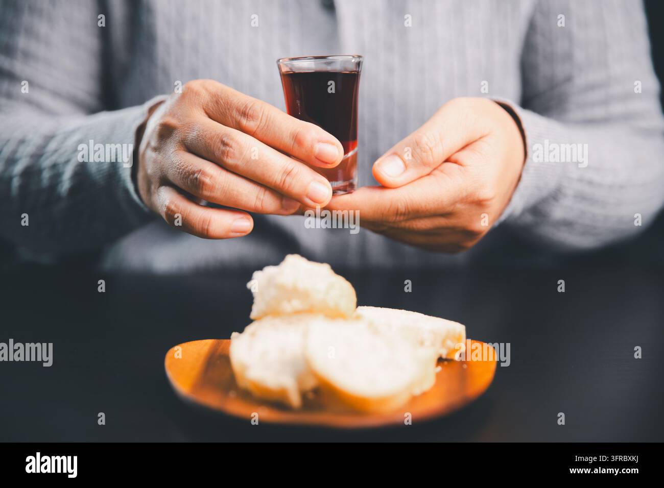 Person tenant un petit verre de boisson de communion rouge foncé au-dessus de pain tranché sur une assiette en bois, illustrant le culte chrétien, le souvenir sacré, Banque D'Images