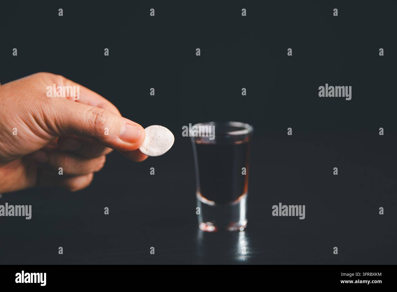 Main tenant une hostie de communion blanche à côté d'un petit verre de boisson rouge foncé sur une table noire, illustrant le culte chrétien, le souvenir sacré, f Banque D'Images