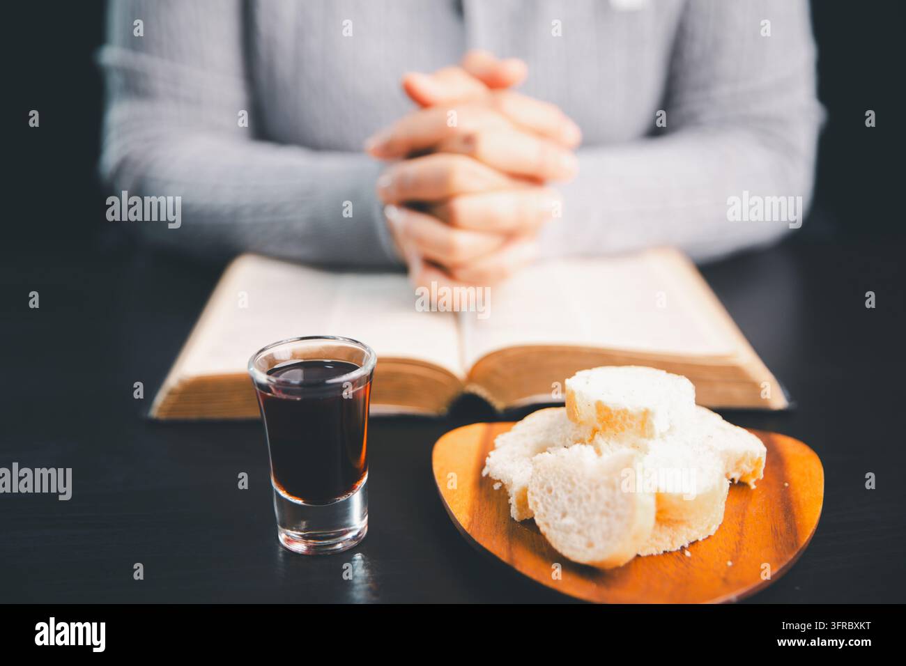 Person en prière derrière un livre ouvert, une boisson de communion rouge foncé et du pain sur une assiette en bois, créant une scène de culte chrétien réfléchie pour la foi Banque D'Images
