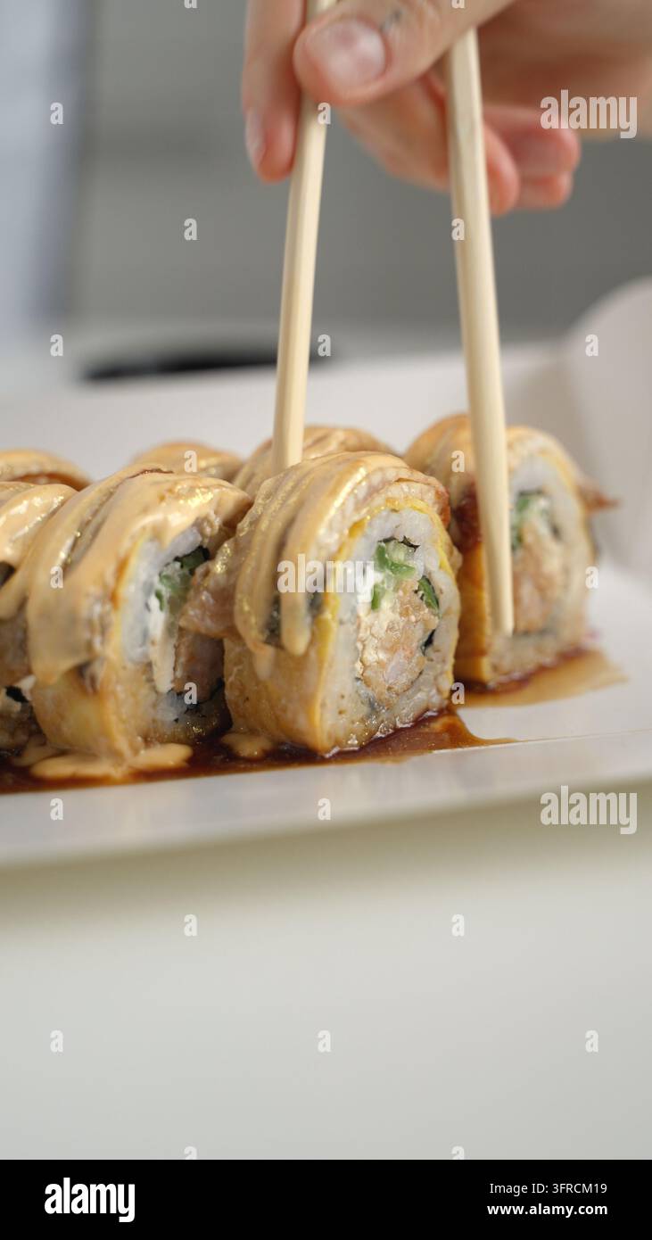 Main utilisant des baguettes pour attraper une délicieuse roulée de sushi saucée d'une assiette blanche, savourant la cuisine asiatique Banque D'Images