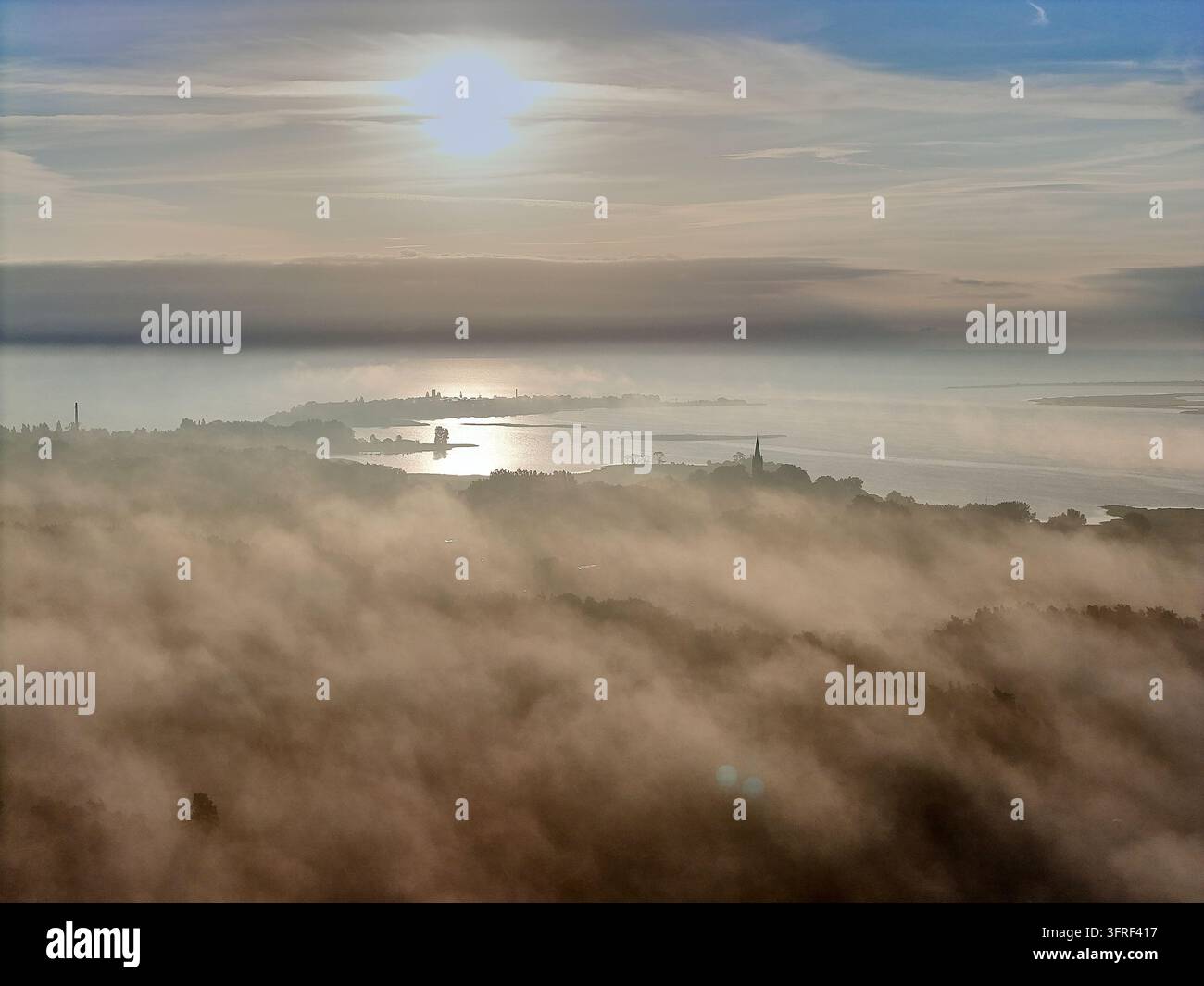 Une vue aérienne éthérée d'un paysage côtier voilé par un épais brouillard matinal, avec le reflet du soleil scintillant sur les eaux calmes de la baie. Silhouette de ville lointaine Banque D'Images