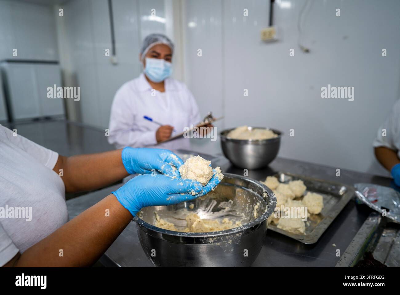 Ouvrier d'usine alimentaire façonnant un mélange de maïs à la main tandis qu'un inspecteur masqué enregistre les données de contrôle qualité dans une salle de traitement. Banque D'Images