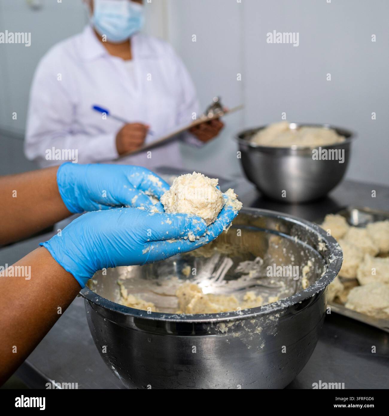 Ouvrier d'usine alimentaire façonnant un mélange de maïs à la main tandis qu'un inspecteur masqué enregistre les données de contrôle qualité dans une salle de traitement. Banque D'Images