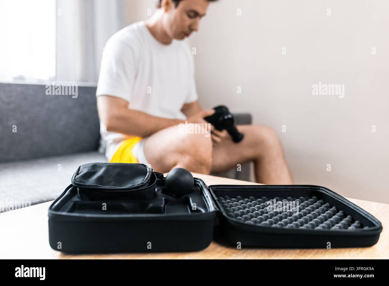 Homme utilisant une masseur électrique sur la jambe à la maison, étui dur ouvert avec accessoires de massage par percussion au premier plan, auto-massage pour les tensions musculaires, après l'entraînement Banque D'Images