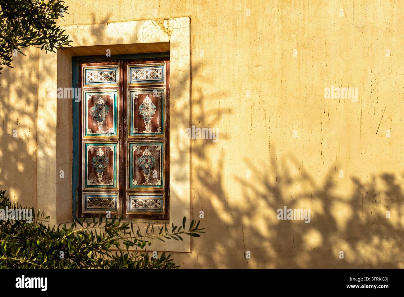 Fenêtre marocaine rustique avec volets en bois peints à la main sur un mur de plâtre jaune à Marrakech, baignée d'une lumière chaude du soleil avec des ombres feuillues. Banque D'Images
