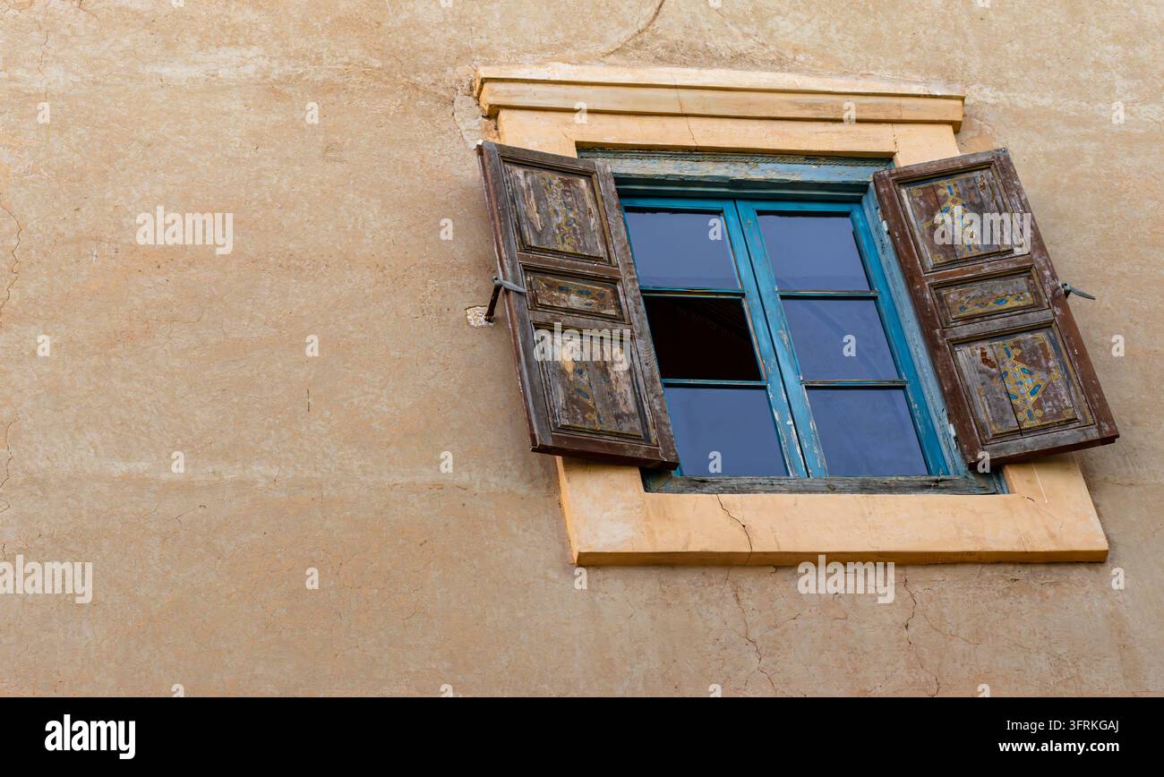 Fenêtre marocaine rustique avec volets en bois peints ouverts sur un mur en adobe ocre texturé, offrant une esthétique architecturale historique et sereine. Banque D'Images