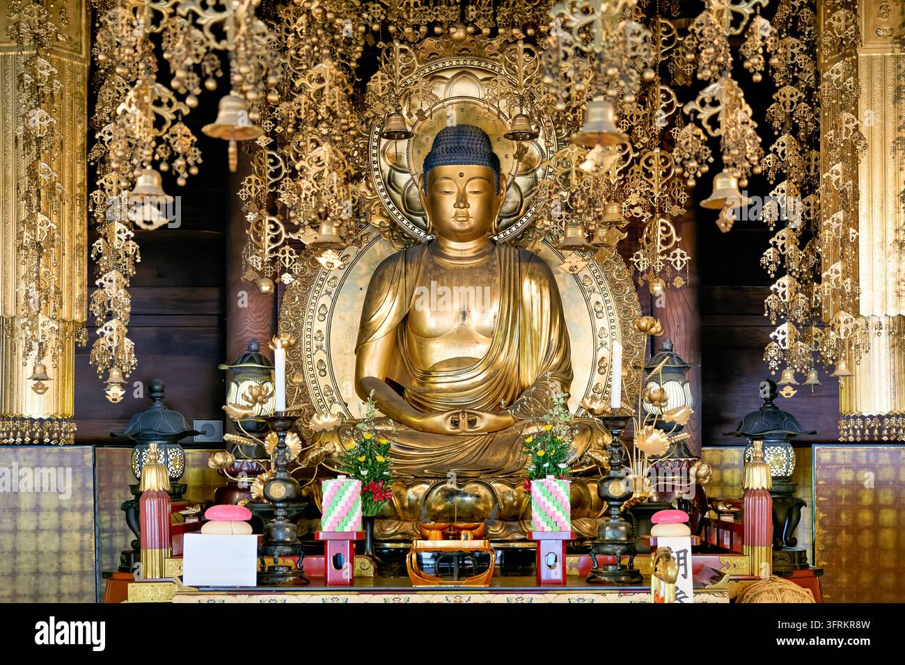 Figure méditante du Bouddha sur un piédestal de lotus avec auréole, encadrée d'ornements suspendus, de fleurs et d'offrandes dans une salle de temple japonais à Kyoto Banque D'Images
