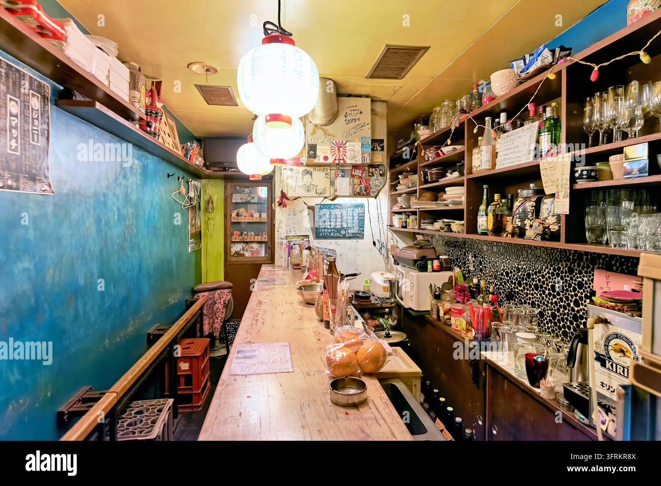Long comptoir en bois bordé de lanternes en papier, étagères de bouteilles d'alcool, verrerie et menus manuscrits à l'intérieur d'un petit lieu de consommation d'alcool au Japon. Banque D'Images